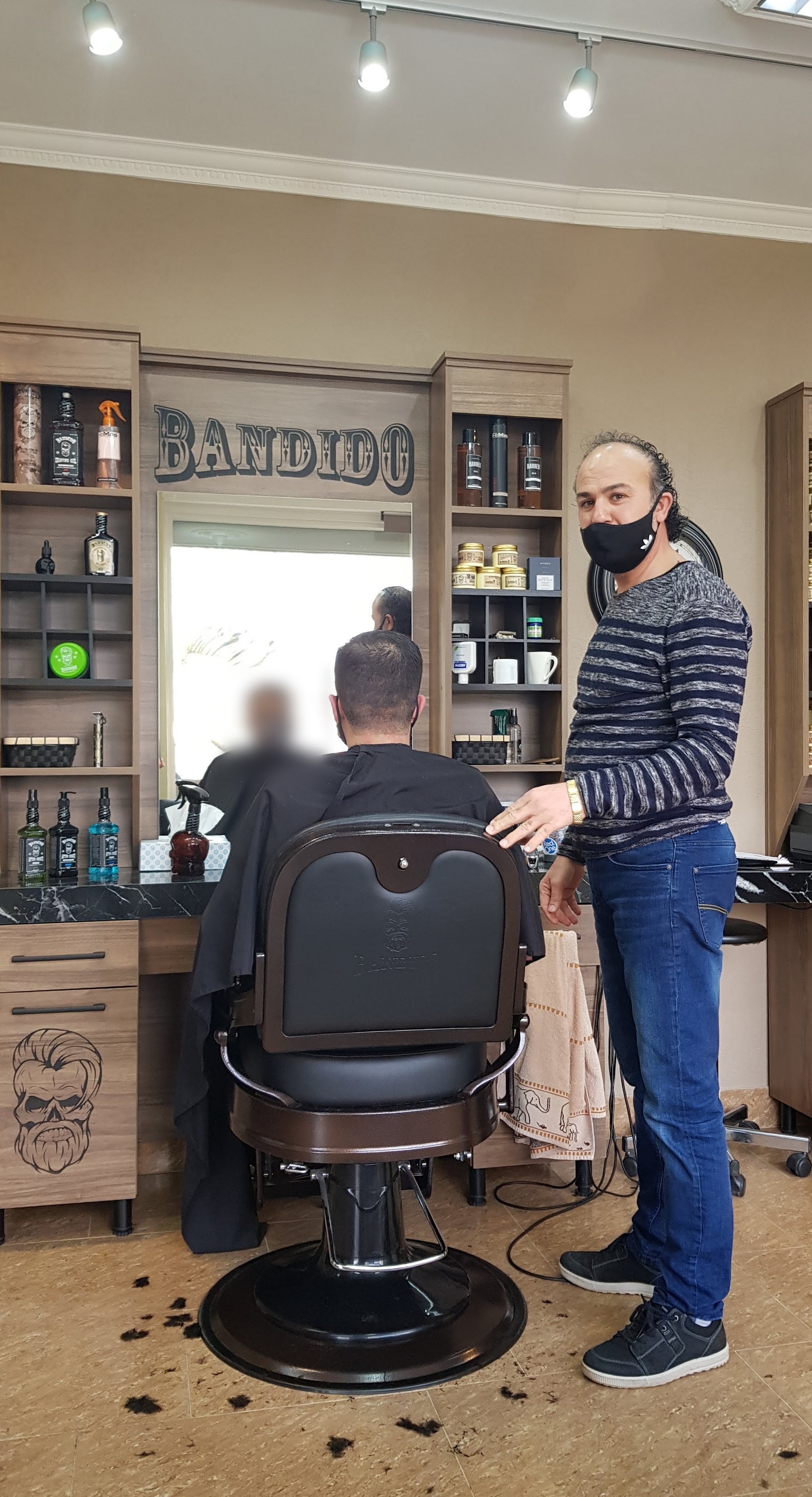 De kapper staat naast een klant in de zwarte kappersstoel, met achter hen de spiegel en de wandkast met Bandido-producten